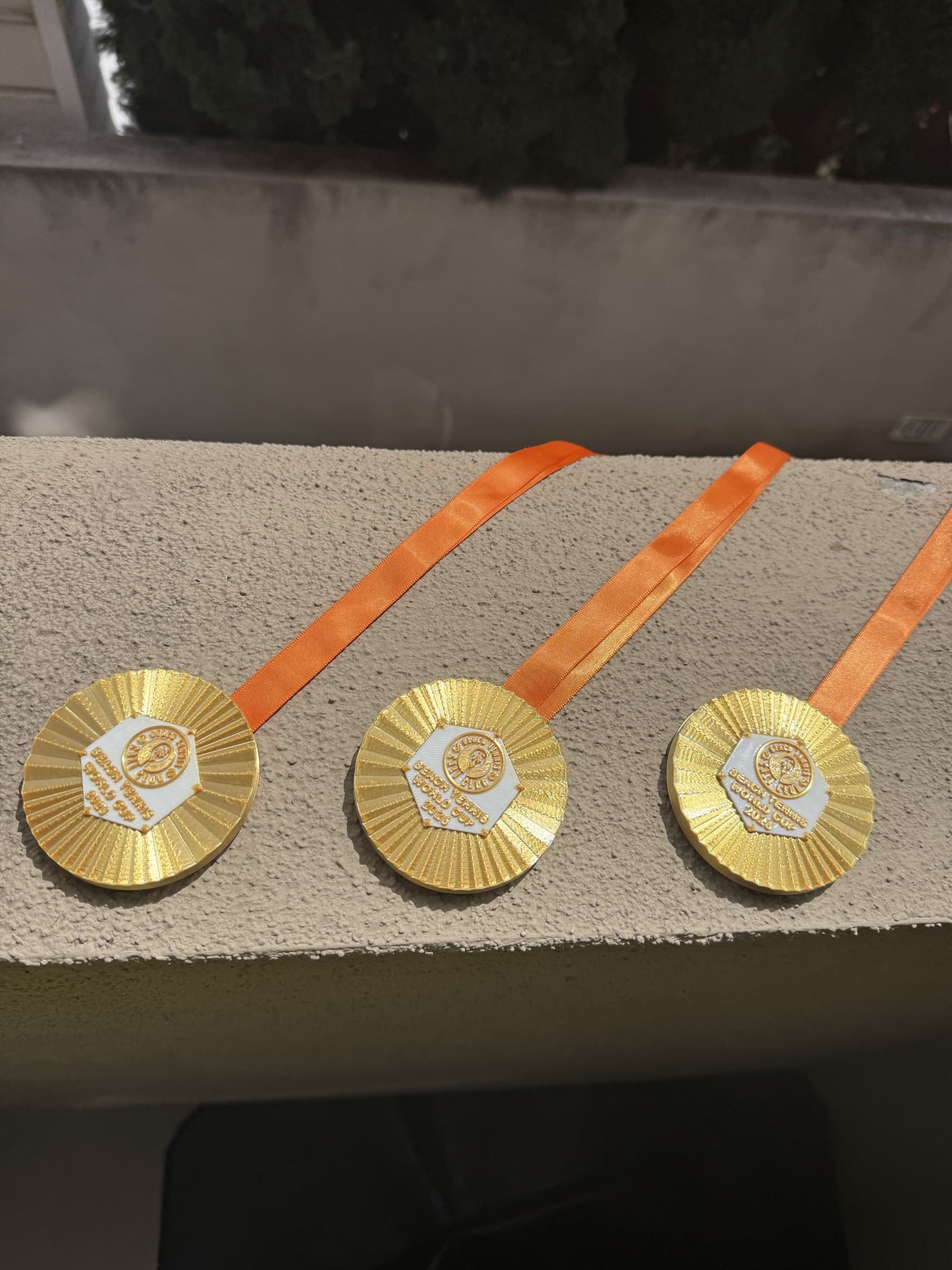 Three custom beach tennis medals with orange ribbons on a ledge